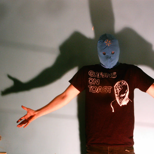 Zainy wearing a t-shirt with 'Greens on Toast' text and a blue gimp mask, casting a shadow on a light background